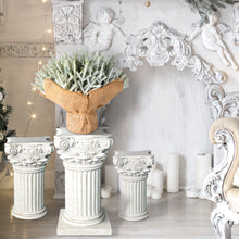 Load image into Gallery viewer, Decorative setting with white columns, a woven basket with Christmas greenery, and ornate wall decorations.
